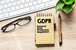 Notesblok med teksten "GDPR General Data Protection Regulation" sidder ved siden af et tastatur, briller, kuglepen og plante på et træskrivebord – ideel som en GDPR skabelon for små virksomheder.