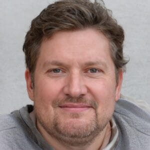 En midaldrende mand med kort, krøllet brunt hår og skæg, iført en lysegrå sweatshirt, smiler let, mens han ser på kameraet mod en neutral baggrund.