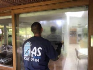 En person i en blå T-shirt renser en stor glasskydedør med en gummiskraber, inde i et rum med et spisebord og stole synligt gennem glasset.