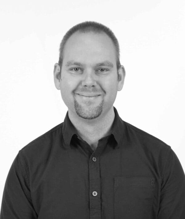 En person med kort hår og fipskæg, iført en knappet skjorte, smiler til kameraet på et sort/hvidt billede.