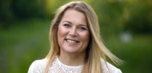 En person med langt blondt hår iført en hvid blondetop smiler udendørs med en sløret grøn baggrund.