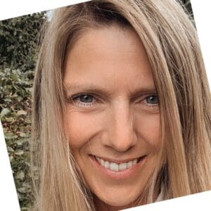 Smilende person med langt blondt hår udendørs, vinklet hovedskud, løv i baggrunden.