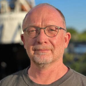 En mand med briller og skæg står udendørs. Han er iført en grå skjorte, og baggrunden er sløret med træer og en synlig bygning.