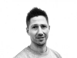 En person med kort hår og en lys sweater smiler let mod en hvid baggrund.